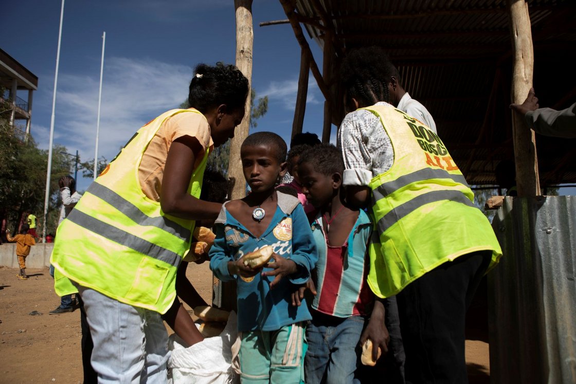 Kinder erhalten Brot von Erwachsenen in 'Don Bosco'-Warnwesten während einer Lebensmittelverteilung.