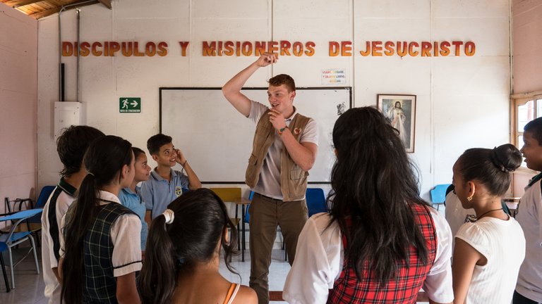 Ein Mann lehrt eine Gruppe aufmerksamer Schüler in einem Raum mit der Aufschrift 'DISCIPULOS Y MISIONEROS DE JESUCRISTO'.