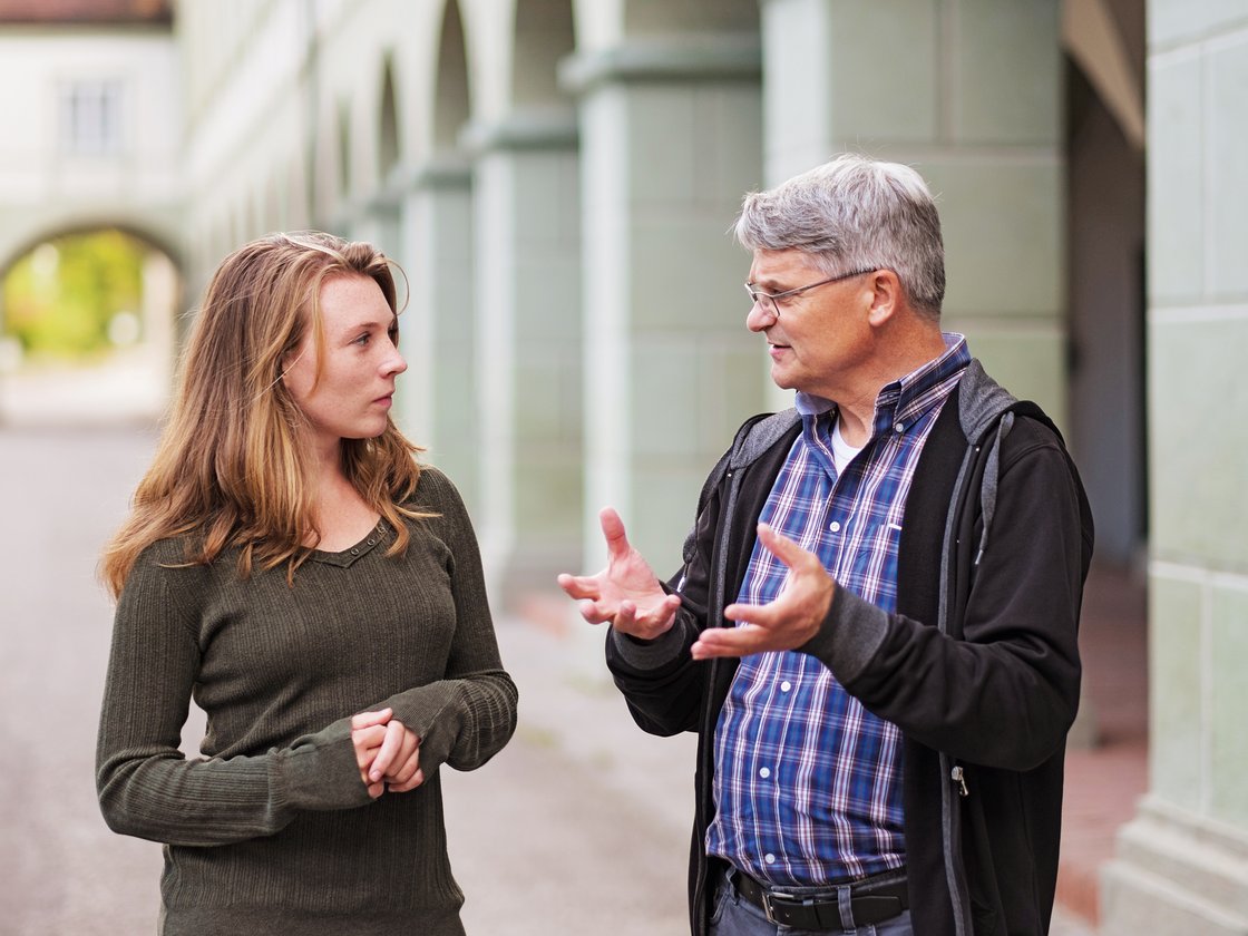 Älterer Mann mit Brille und jüngere Frau im ernsten Gespräch im Freien, Bögen im Hintergrund.
