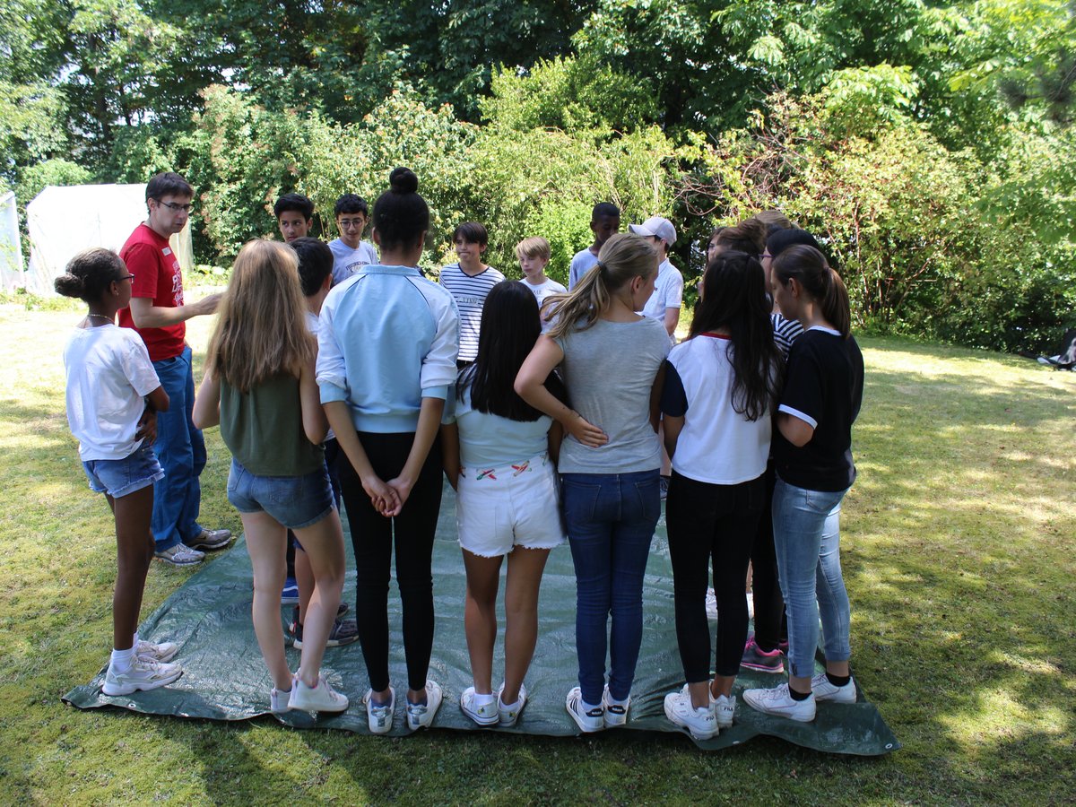 Eine Gruppe Jugendlicher steht im Kreis auf einer grünen Plane im Gras, umgeben von Bäumen, vermutlich eine Outdoor-Aktivität.