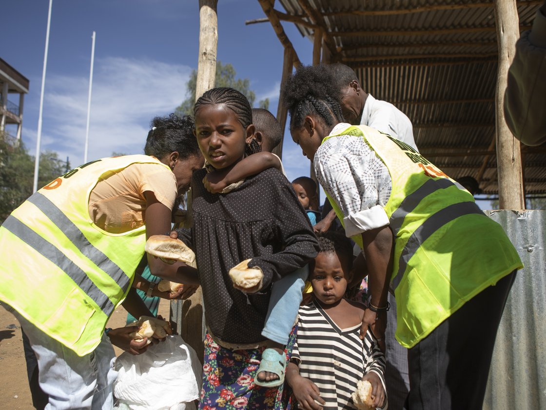 Kinder und Erwachsene, darunter ein Mädchen mit einem Kind, erhalten Brot von Helfern in gelben Westen im Freien.