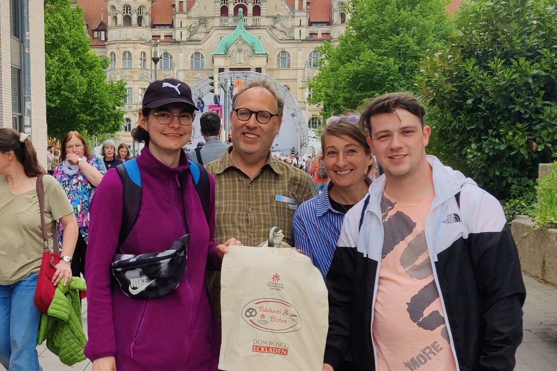 Vier lächelnde Personen posieren draußen vor einem großen historischen Gebäude mit grünen Dächern; eine hält eine 'Don Bosco'-Tüte.