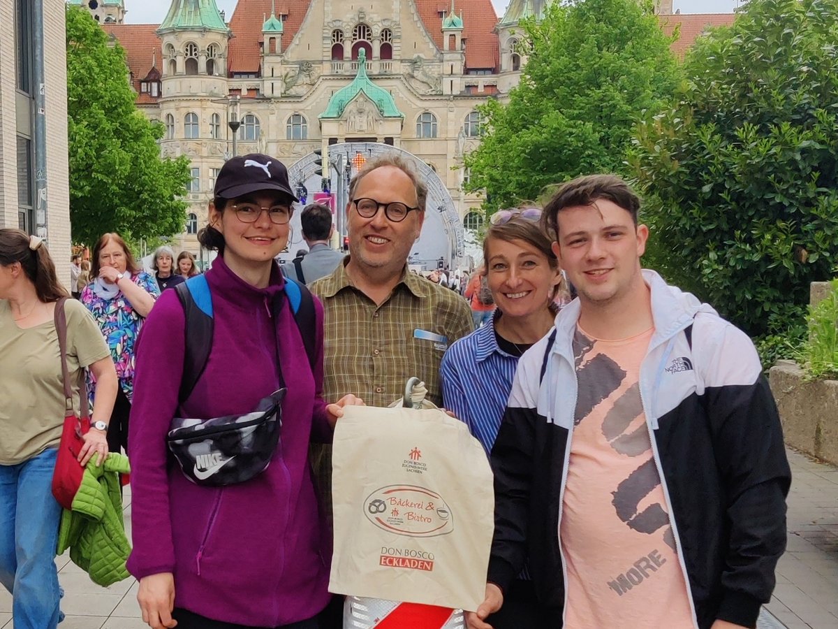 Vier lächelnde Personen posieren draußen vor einem großen historischen Gebäude mit grünen Dächern; eine hält eine 'Don Bosco'-Tüte.