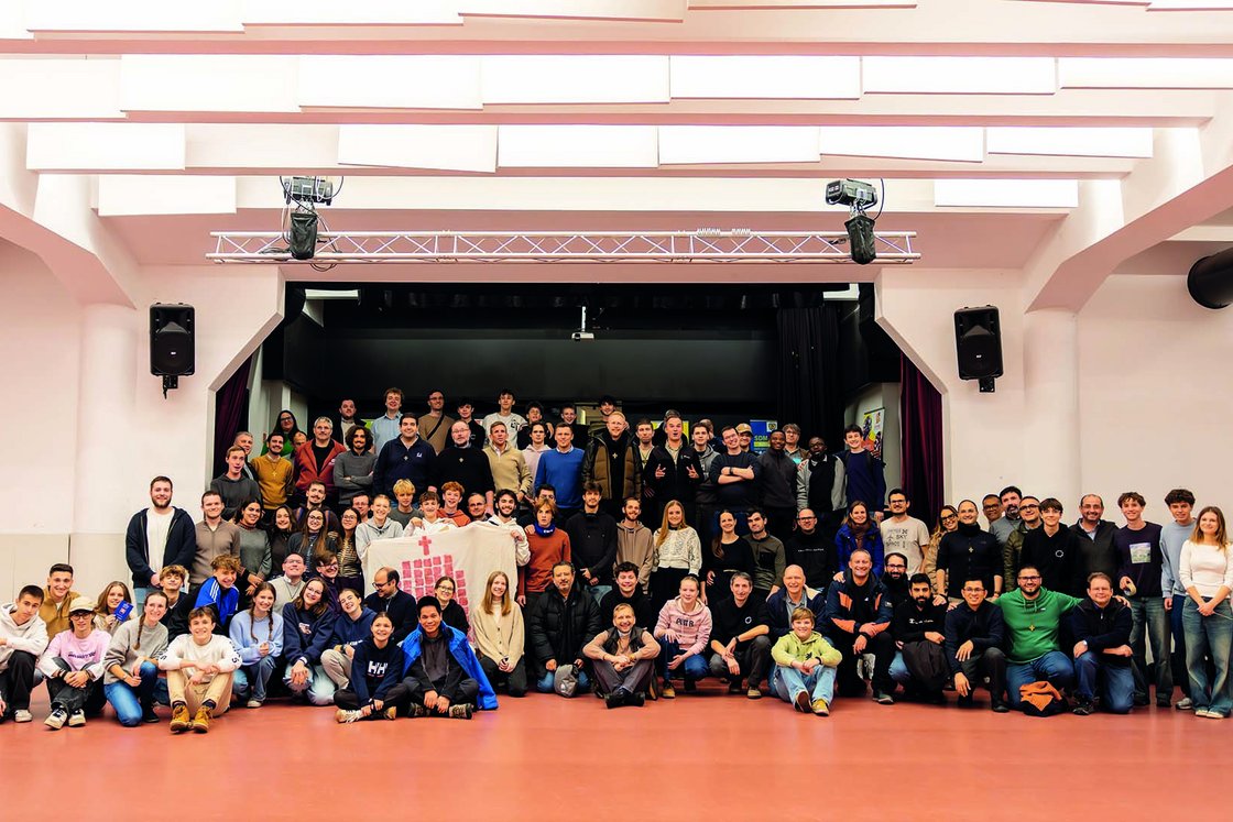Große Gruppe lächelnder Menschen jeden Alters in einer Halle, die ein weißes Banner mit einem roten Kreuz hält, Bühne im Hintergrund.