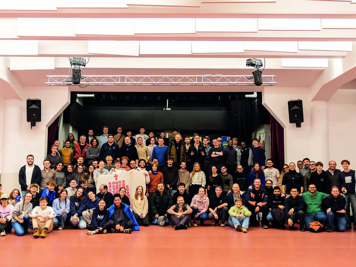 Große Gruppe lächelnder Menschen jeden Alters in einer Halle, die ein weißes Banner mit einem roten Kreuz hält, Bühne im Hintergrund.