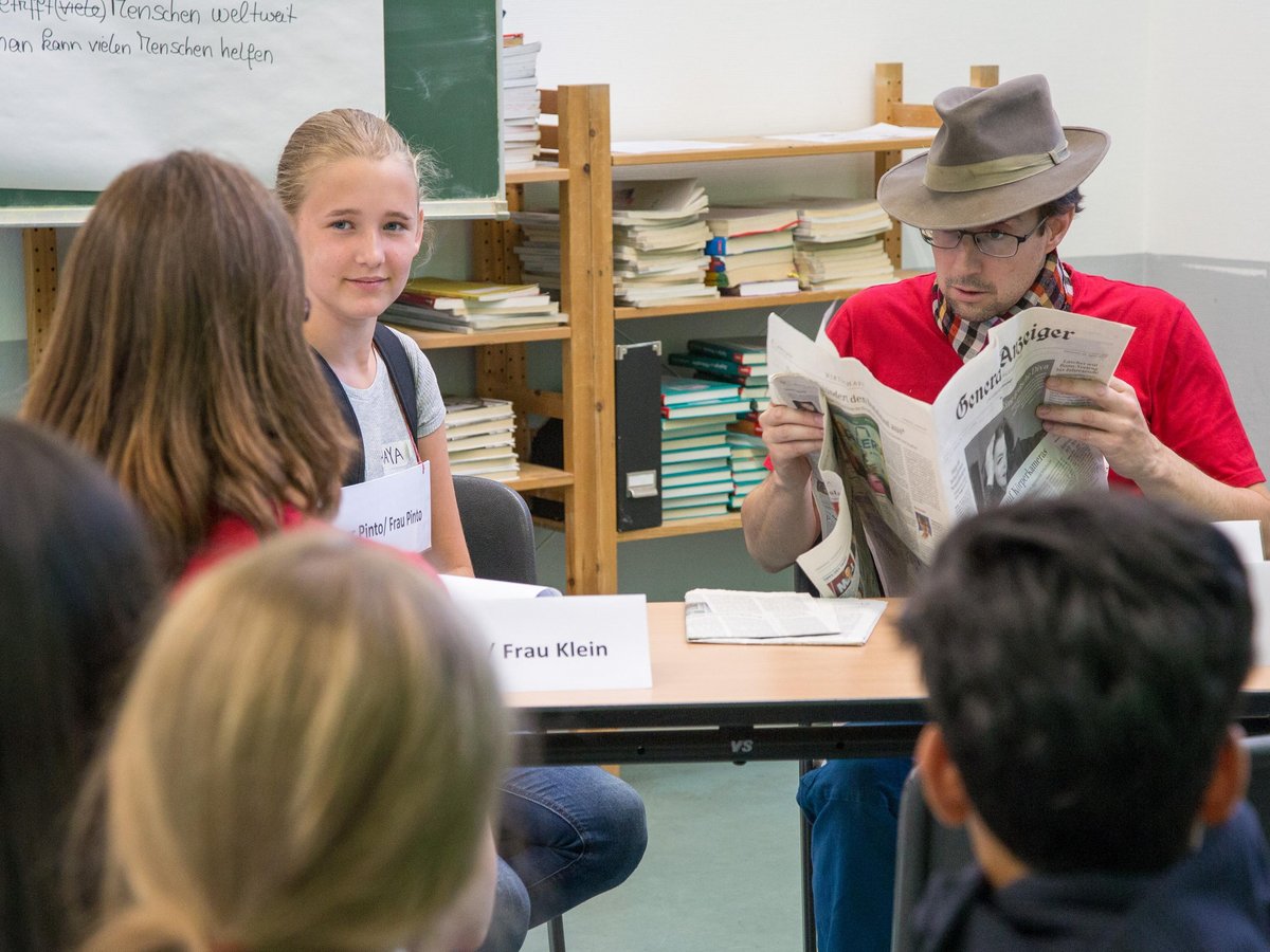 Ein Mann mit Hut liest Zeitung, ein junges Mädchen lächelt, und Schüler nehmen an einem Klassenzimmer-Workshop teil.