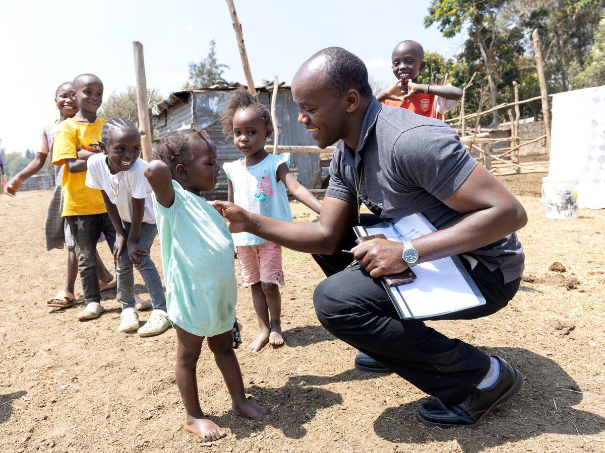 Ein lächelnder Mann hockt, spricht mit einem kleinen Mädchen, umgeben von anderen fröhlichen Kindern in ländlicher Gegend.