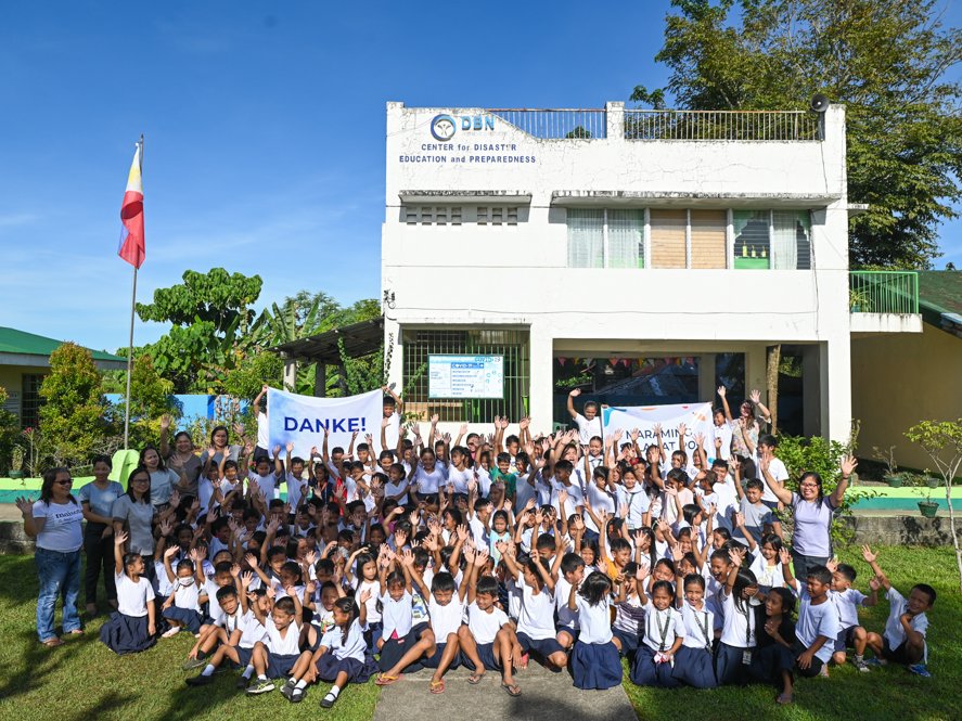 Eine große Gruppe philippinischer Kinder und Erwachsener winkt fröhlich vor dem Katastrophenschutz-Zentrum mit einem „DANKE!“-Banner.