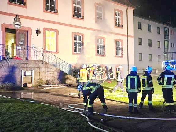 Feuerwehrleute bekämpfen nachts einen Brand in einem Gebäude, Rauch steigt auf, Schläuche liegen auf dem Rasen.