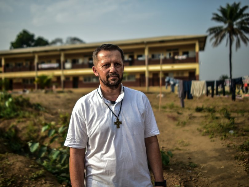 Ein bärtiger Mann in einem weißen Hemd mit Kreuzkette steht vor einem mehrstöckigen Gebäude, Palme und Wäscheleine im Hintergrund.