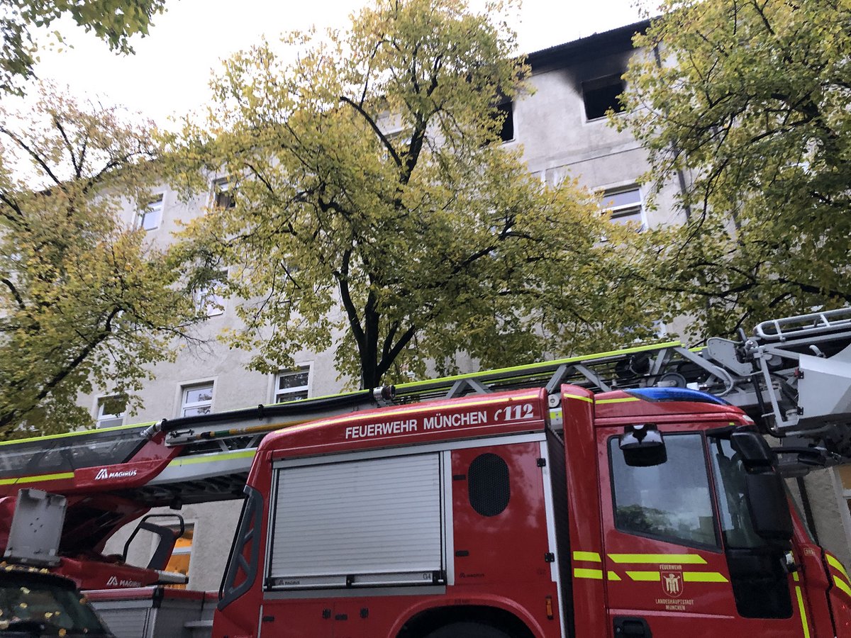 Rotes Feuerwehrauto der "Feuerwehr München" mit ausgefahrener Leiter vor einem Gebäude mit Brandschäden im Obergeschoss.