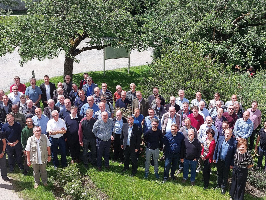 Eine große Gruppe von hauptsächlich Männern und einigen Frauen posiert für ein Gruppenfoto auf einer grünen Wiese unter Bäumen.