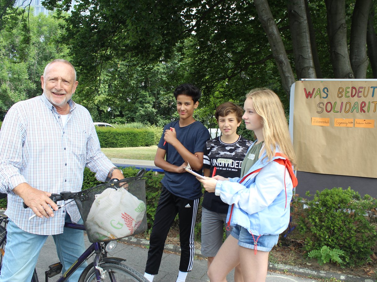 Ein älterer Mann mit Fahrrad und drei Jugendlichen vor einem Schild, das nach der Bedeutung von Solidarität fragt.