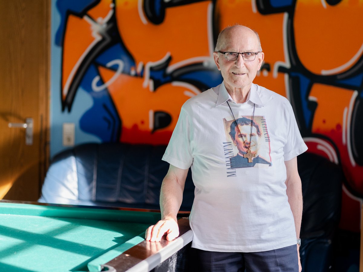 Lächelnder älterer Mann mit Brille in einem Marianum Buxheim T-Shirt und Kreuz, steht am Billardtisch vor Graffiti.