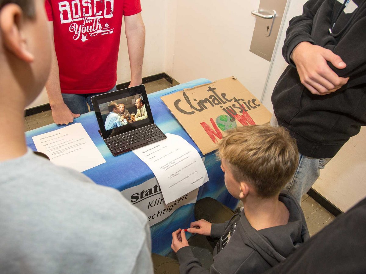 Schüler an einer Klimagerechtigkeits-Station mit Laptop (Video), Papieren und einem 'Climate justice NOW'-Schild.