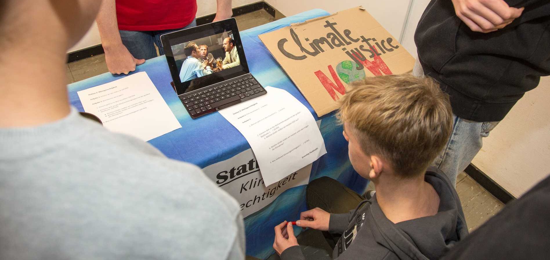 Schüler an einer Klimagerechtigkeits-Station mit Laptop (Video), Papieren und einem 'Climate justice NOW'-Schild.