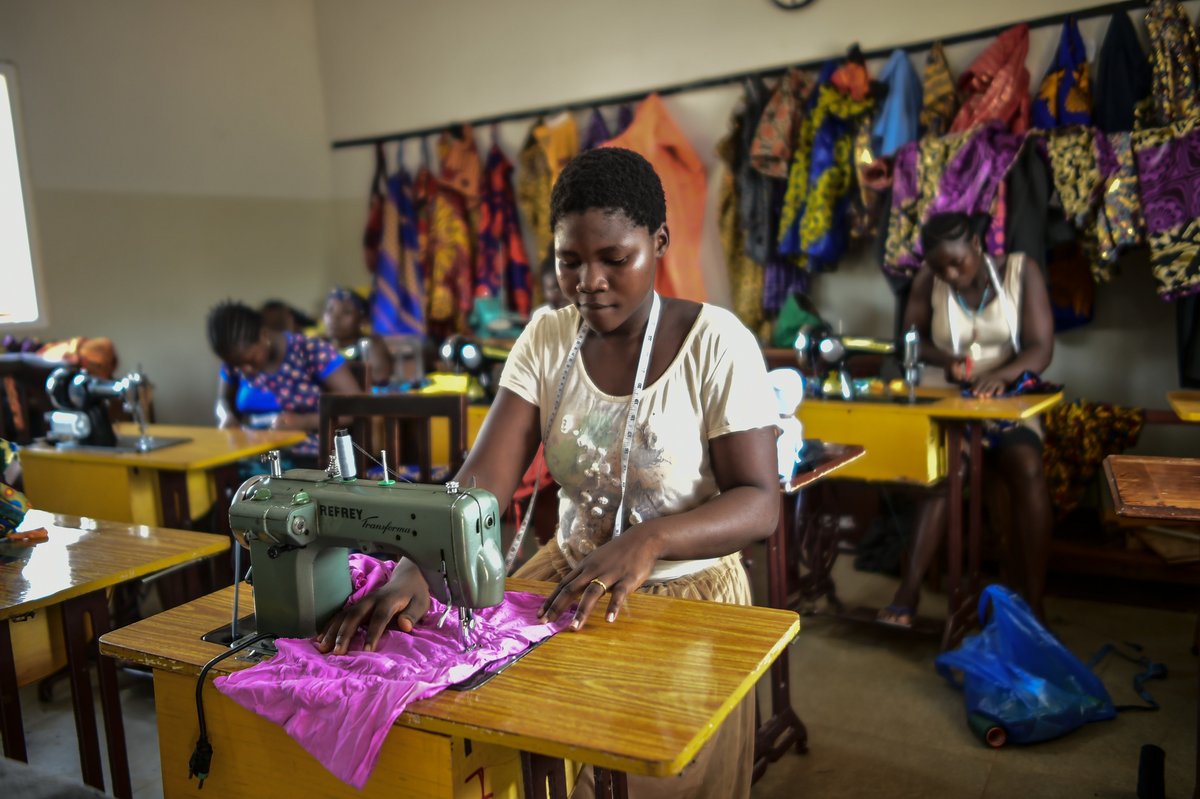 Eine junge Frau näht mit einer Nähmaschine pinkfarbenen Stoff in einer lebhaften Werkstatt. Andere Frauen und bunte Kleidung sind sichtbar.