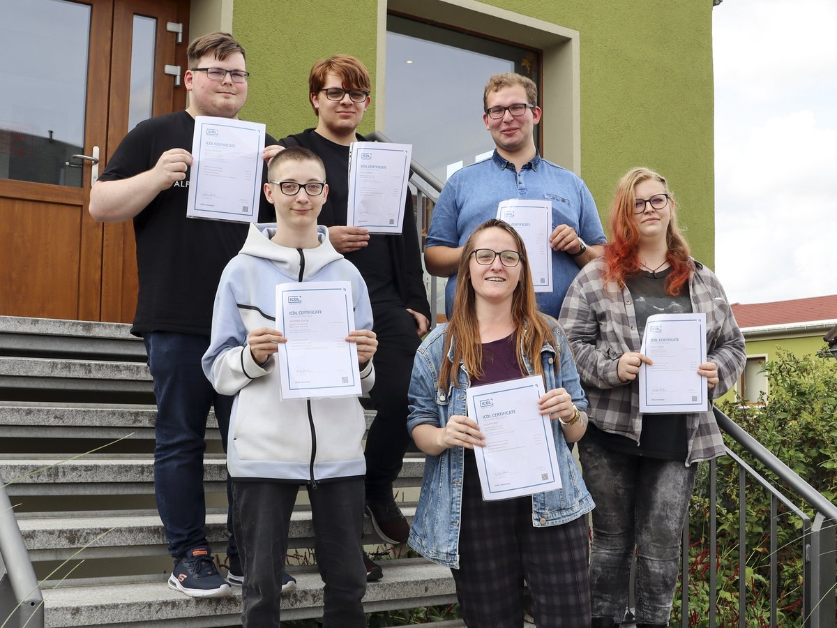 Sieben lächelnde junge Leute auf einer Treppe präsentieren stolz ihre ICDL-Zertifikate, eine Computerkenntnis-Auszeichnung.