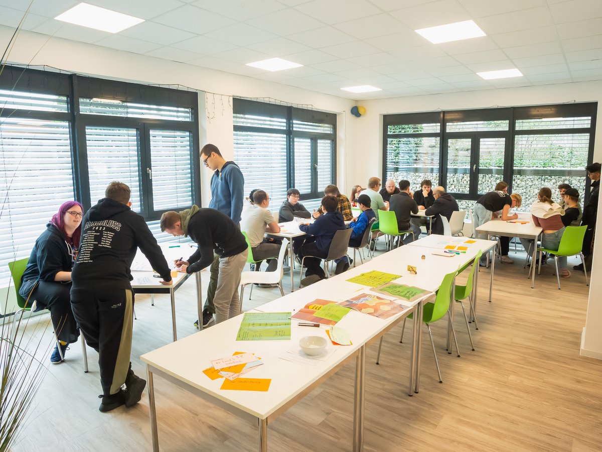 Schüler arbeiten in Gruppen an Tischen in einem hellen Klassenzimmer oder Workshop.