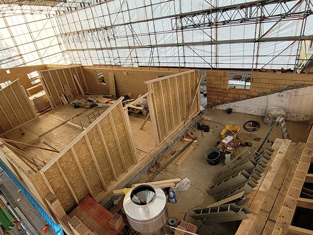 Innenansicht einer Baustelle mit Holzständerwänden und einer teilweise gemauerten Wand unter einem provisorischen Gerüstdach.