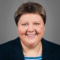 Porträt einer lächelnden Frau mit kurzen braunen Haaren, blauen Augen, Perlenohrring, dunklem Blazer, gestreiftem Oberteil und Silberkette.