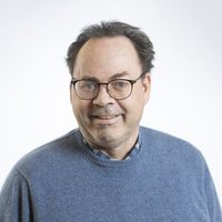 Ein lächelnder Mann mit Brille, grauem Haar und blauem Pullover posiert für ein Porträt vor weißem Hintergrund.