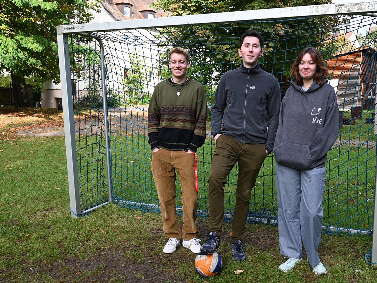 Drei junge Leute posieren vor einem Fußballtor mit einem Ball auf einem Rasenplatz.