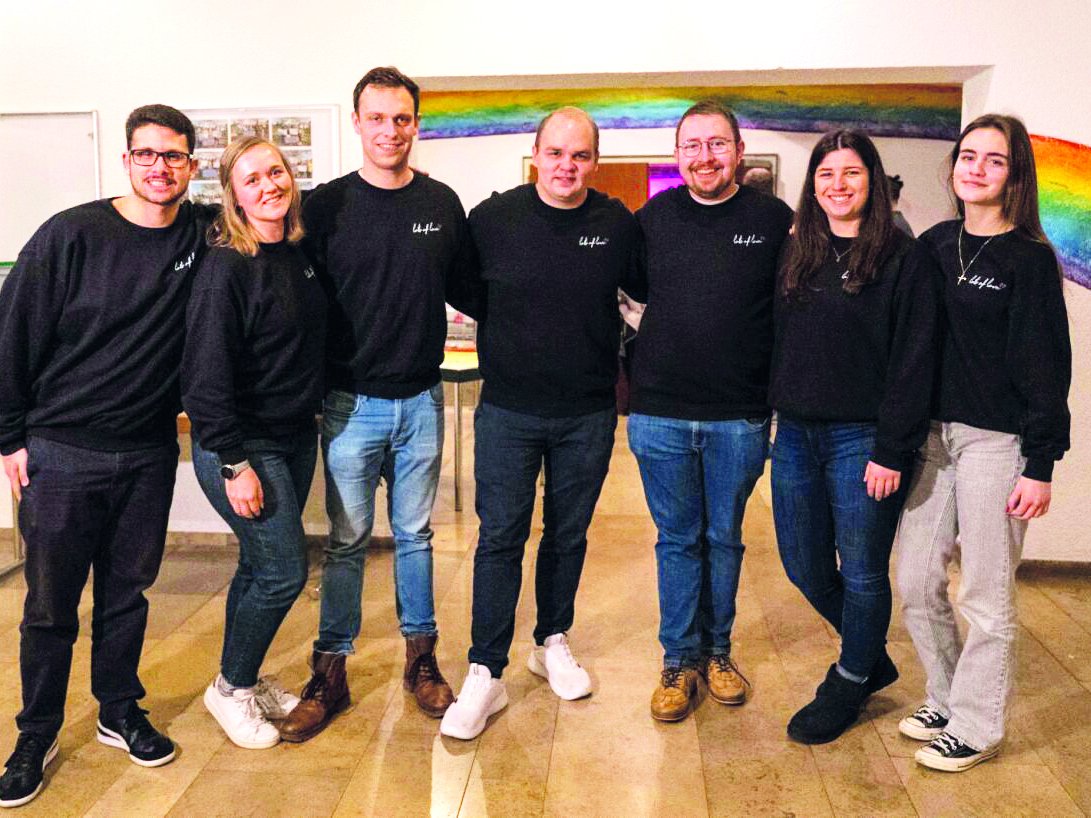 Sieben lächelnde Personen in schwarzen Oberteilen und Jeans stehen vor einer Wand mit Regenbogen.