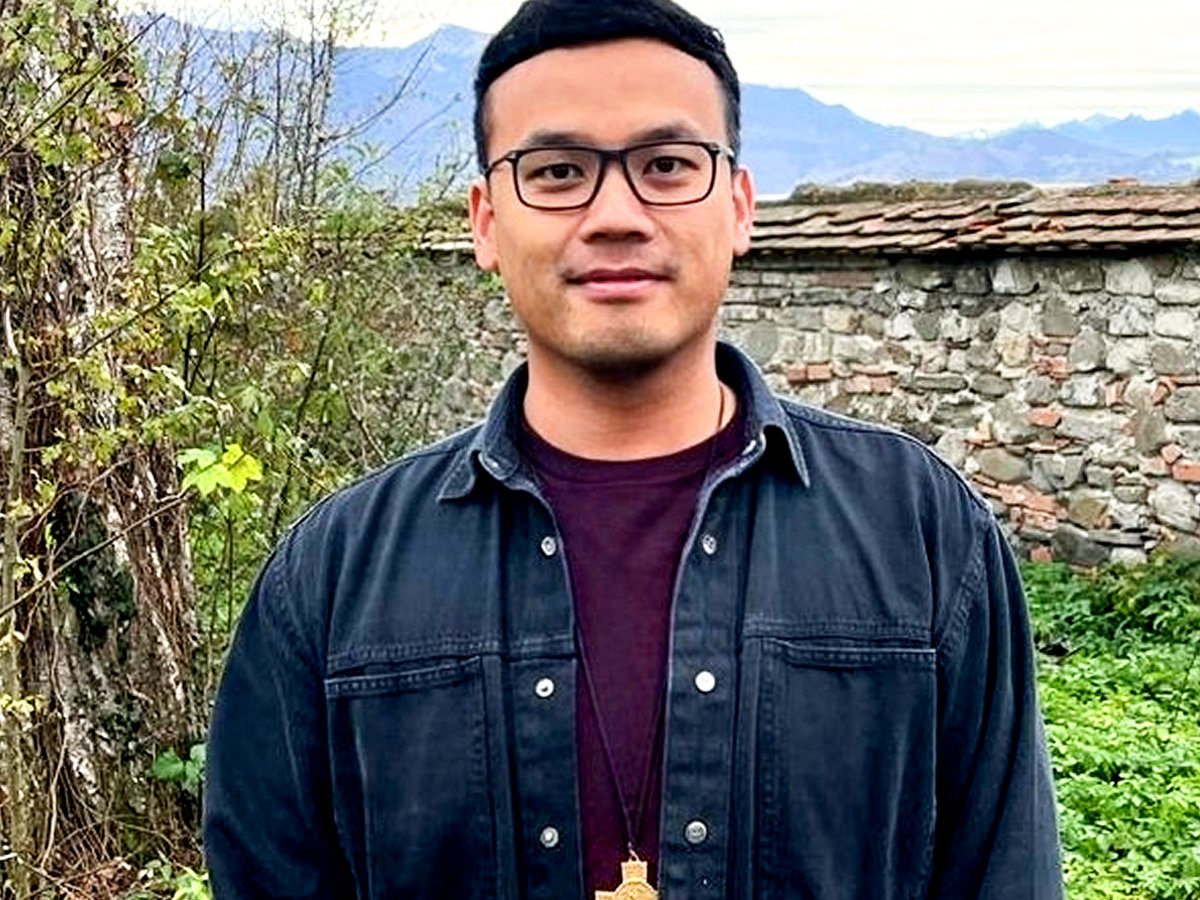 Ein Mann mit Brille und einem goldenen Kreuzanhänger steht draußen vor einer Steinmauer und Bergen.