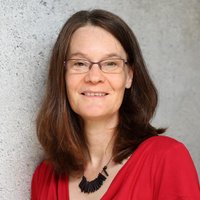 Porträt einer lächelnden Frau mit braunen Haaren, Brille und rotem Oberteil vor einer grauen Wand.