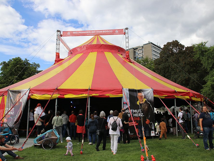 Ein großes rot-gelb gestreiftes Don Bosco Veranstaltungszelt mit eintretenden Personen, auf grünem Rasen unter bewölktem Himmel.