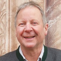 Ein lächelnder Mann mittleren Alters mit grauen Haaren und blauen Augen, trägt eine graue Trachtenjacke mit grünem Kragen.