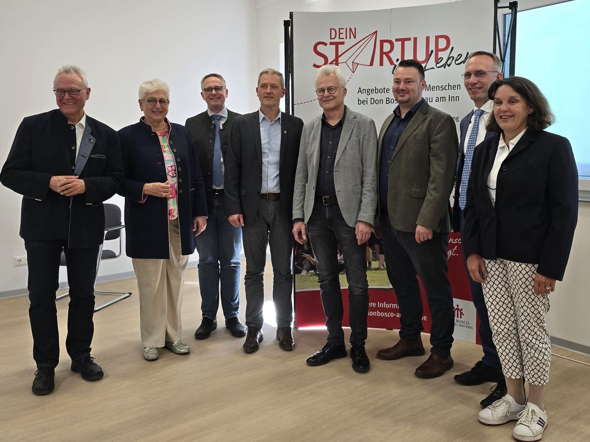 Acht Personen, sechs Männer und zwei Frauen, lächeln vor einem 'DEIN STARTUP LEBEN' Banner bei Don Bosco am Inn.