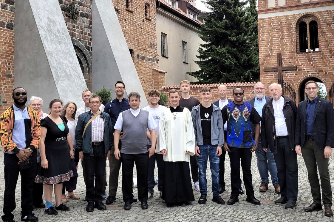 Eine vielfältige Gruppe von Männern und Frauen, darunter Geistliche, steht auf einem Kopfsteinpflasterhof vor alten Backsteingebäuden.