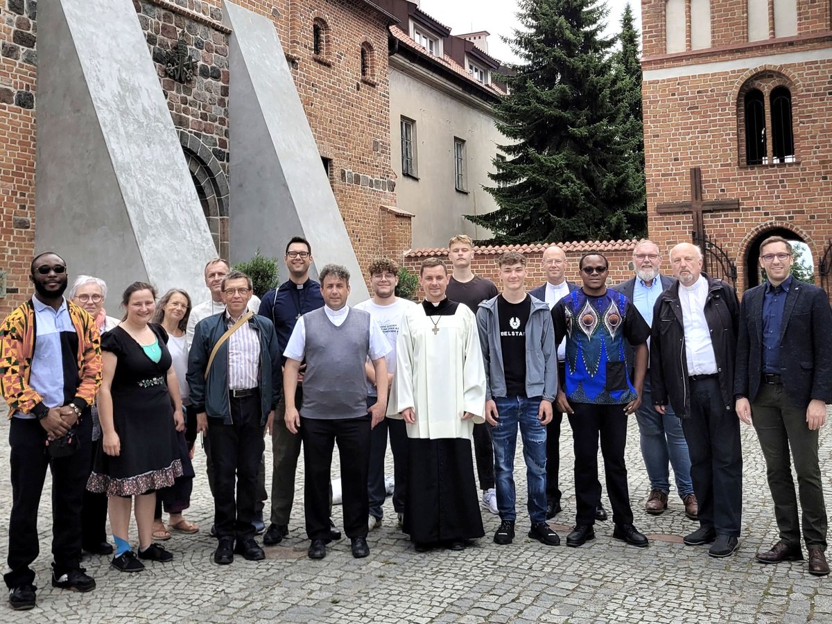 Eine vielfältige Gruppe von Männern und Frauen, darunter Geistliche, steht auf einem Kopfsteinpflasterhof vor alten Backsteingebäuden.