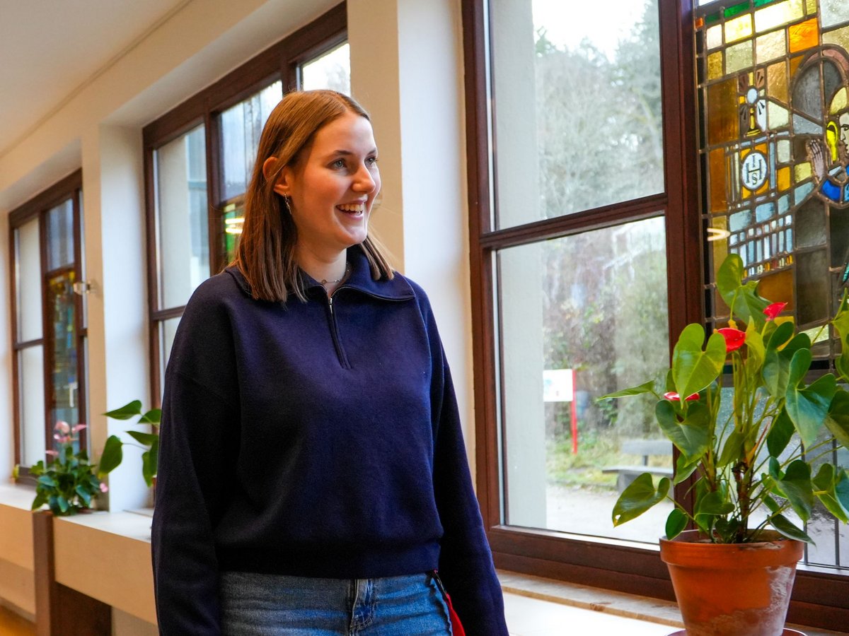 Lächelnde junge Frau in blauem Sweatshirt steht an einem großen Fenster mit Buntglasabschnitt und Topfpflanze.