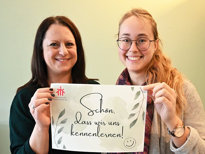 Zwei lächelnde Frauen halten ein Schild mit der Aufschrift 'Schön, dass wir uns kennenlernen!' und dem Salesianer Don Boscos Logo.