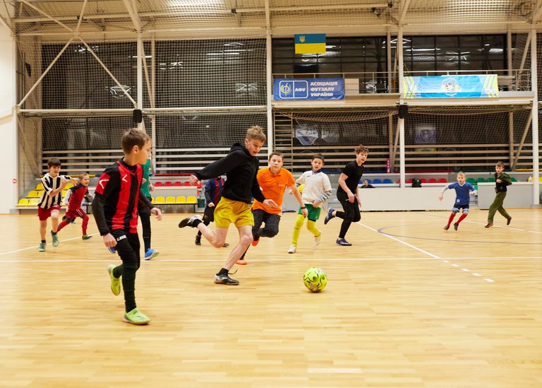 Junge Jungen spielen Futsal auf einem Indoor-Spielfeld mit ukrainischer Flagge und Bannern im Hintergrund.