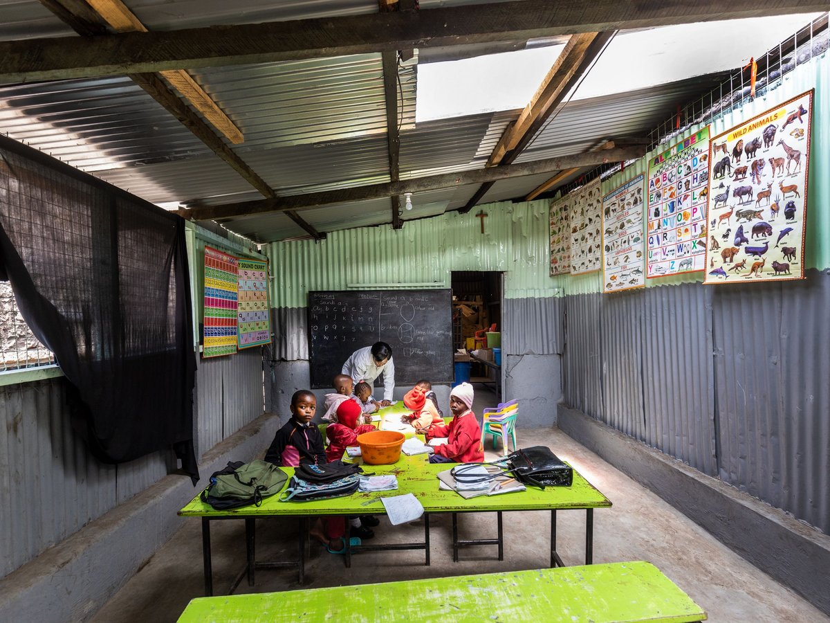 Eine Lehrerin hilft kleinen Kindern an grünen Tischen in einem rustikalen Klassenzimmer mit Wellblechwänden und Lehrpostern.