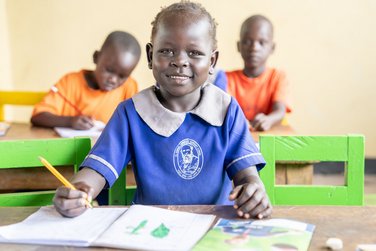 Lächelndes Schulkind in blauer Uniform, hält Bleistift, sitzt am Schreibtisch im Klassenzimmer, andere Kinder im Hintergrund.