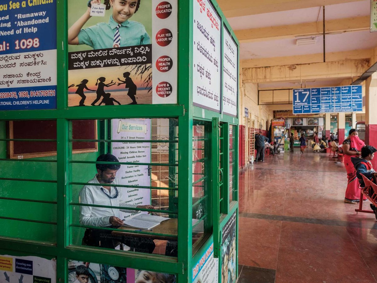 Ein Mann in einem grünen Kinderhilfsstand an einem belebten indischen Busbahnhof. Schilder werben für Kinderrechte und Hilfsnummern.