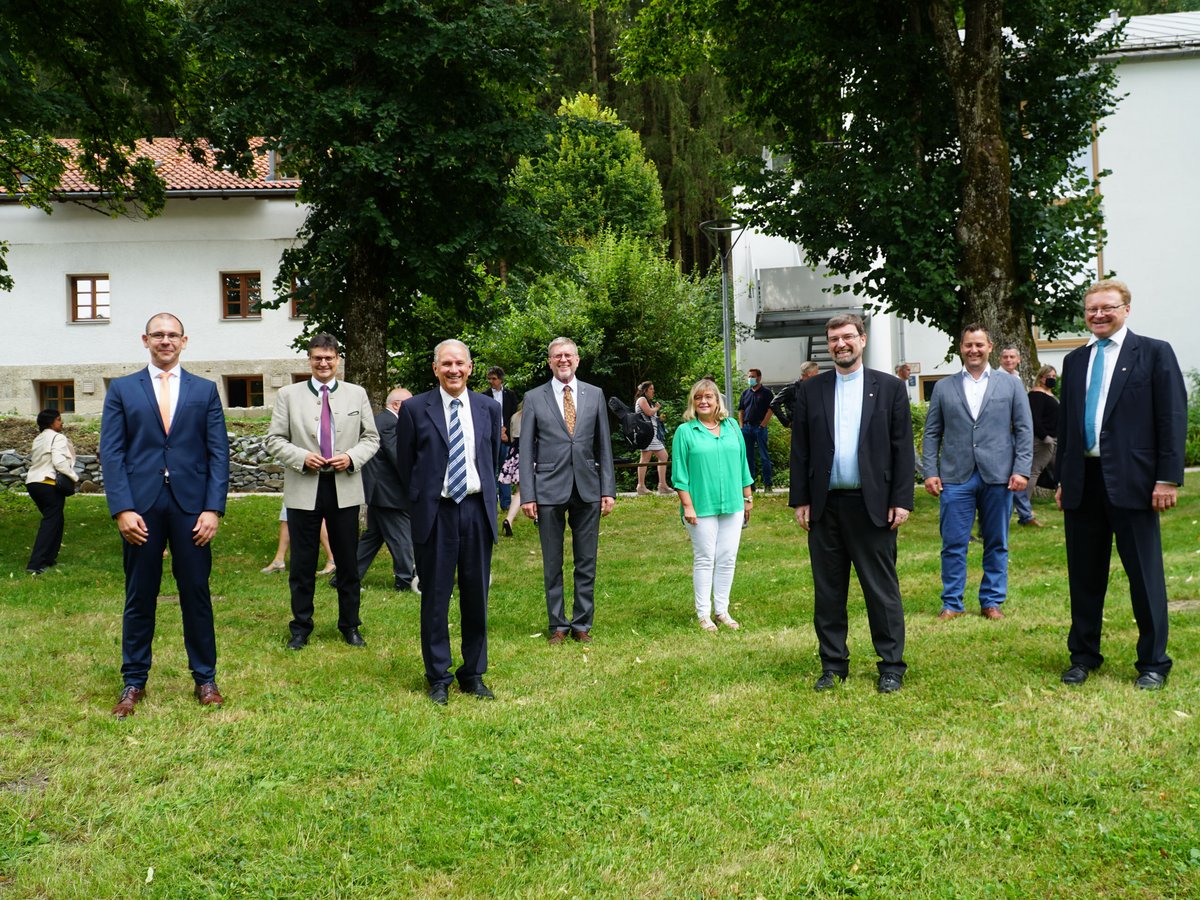 Eine Gruppe formell gekleideter Männer und einer Frau steht lächelnd auf einer Wiese vor Gebäuden und Bäumen.