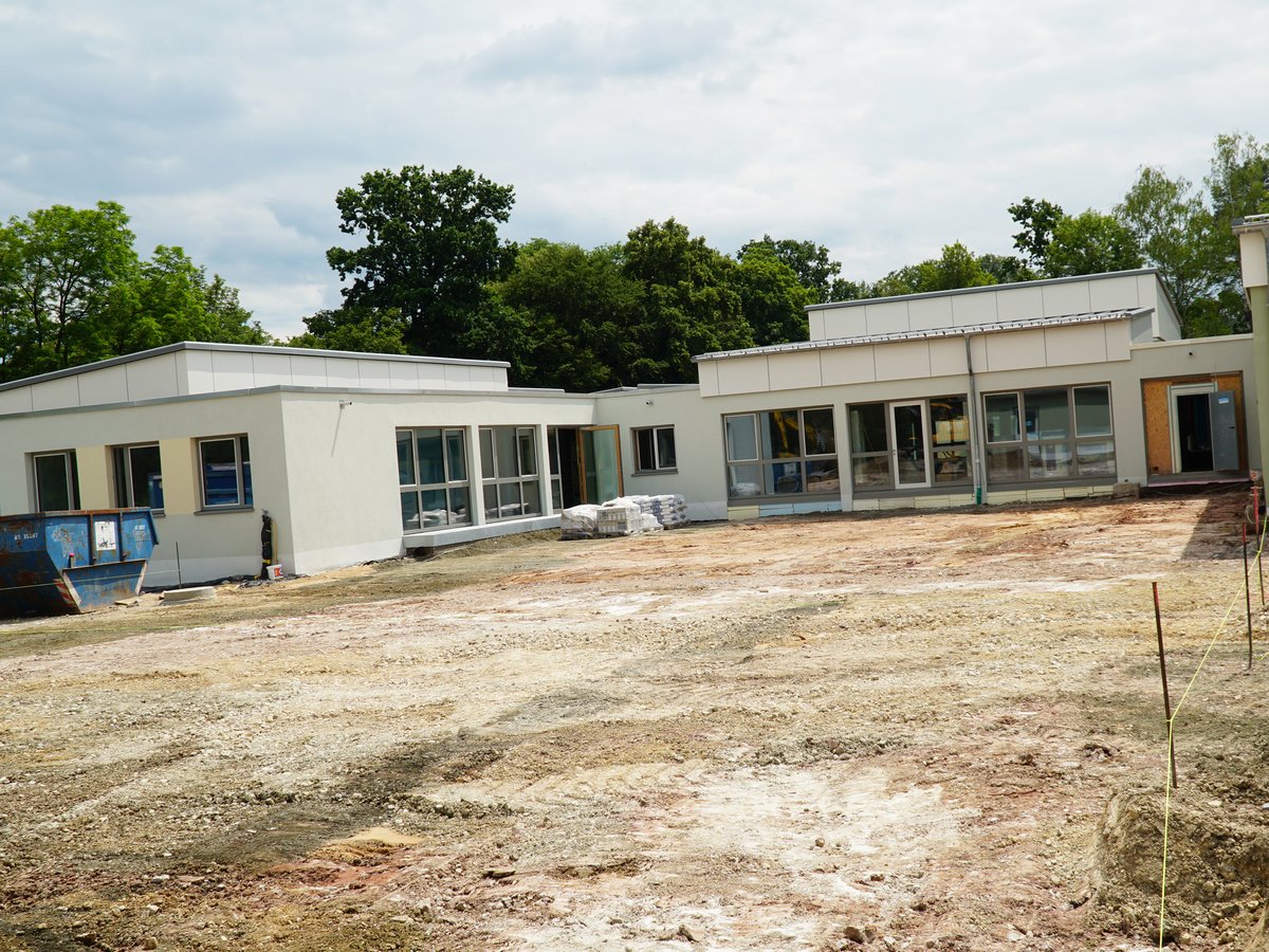 Baustelle mit hellen Modulgebäuden, unbefestigtem Boden, blauem Container und Materialstapeln unter bewölktem Himmel.