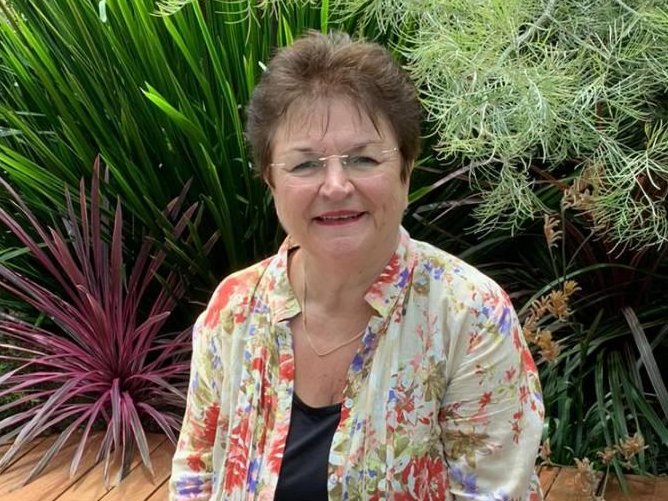 Eine ältere Frau mit Brille und Blumenhemd lächelt auf einer Holzbank inmitten üppiger grüner Gartenpflanzen.