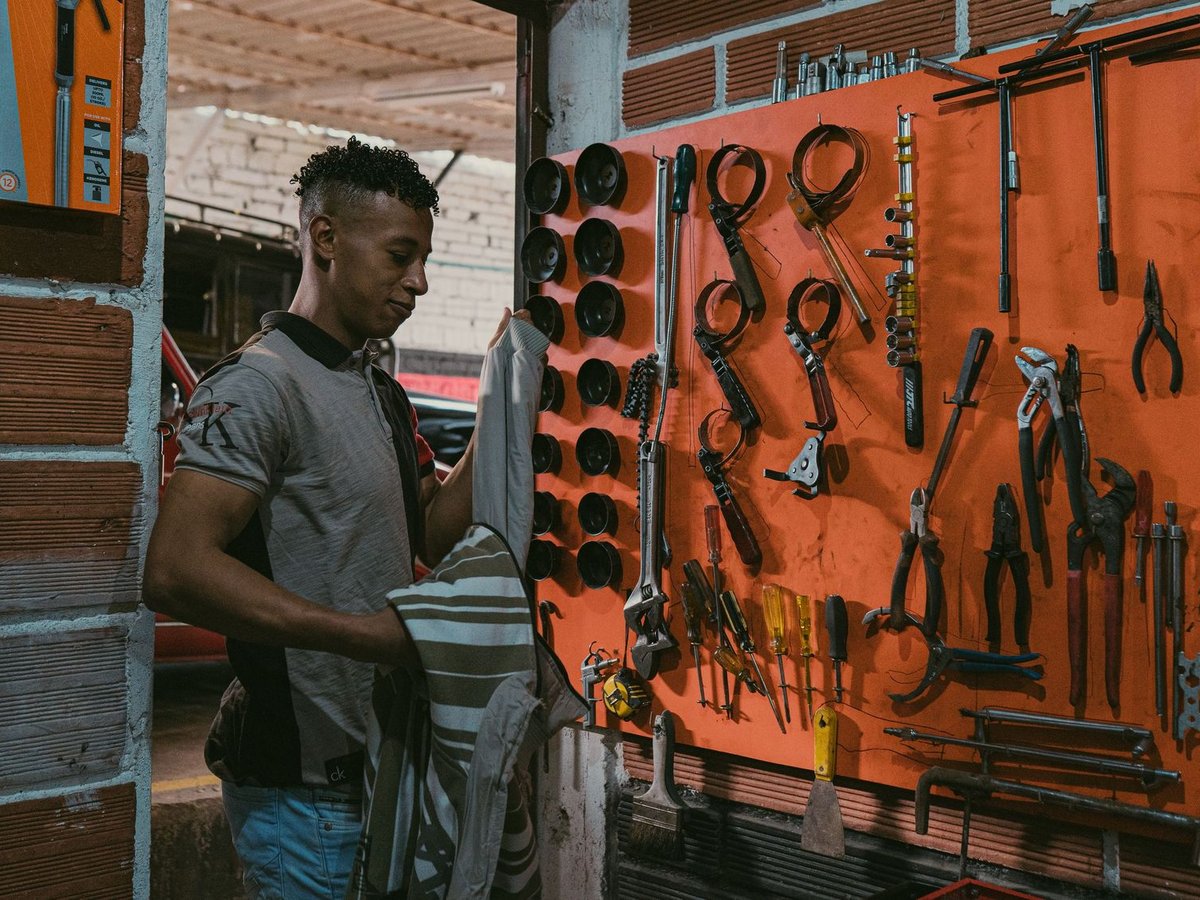 Junger Mann in einer Werkstatt mit oranger Werkzeugwand, hält ein gestreiftes Kleidungsstück, bereit zur Arbeit.