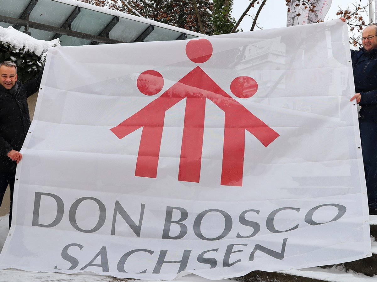 Zwei lächelnde Männer halten ein weißes Banner mit rotem Logo (drei Figuren/Haus) und dem Text "DON BOSCO SACHSEN" im Schnee.