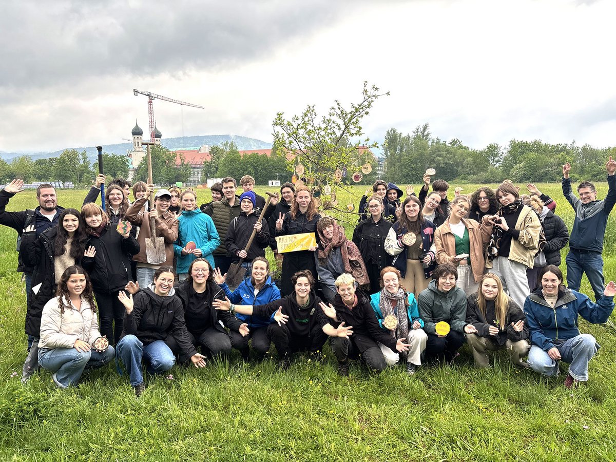 Eine vielfältige Gruppe von Menschen posiert auf einer Wiese mit einem geschmückten Baum und Werkzeugen. Kloster und Kran im Hintergrund.
