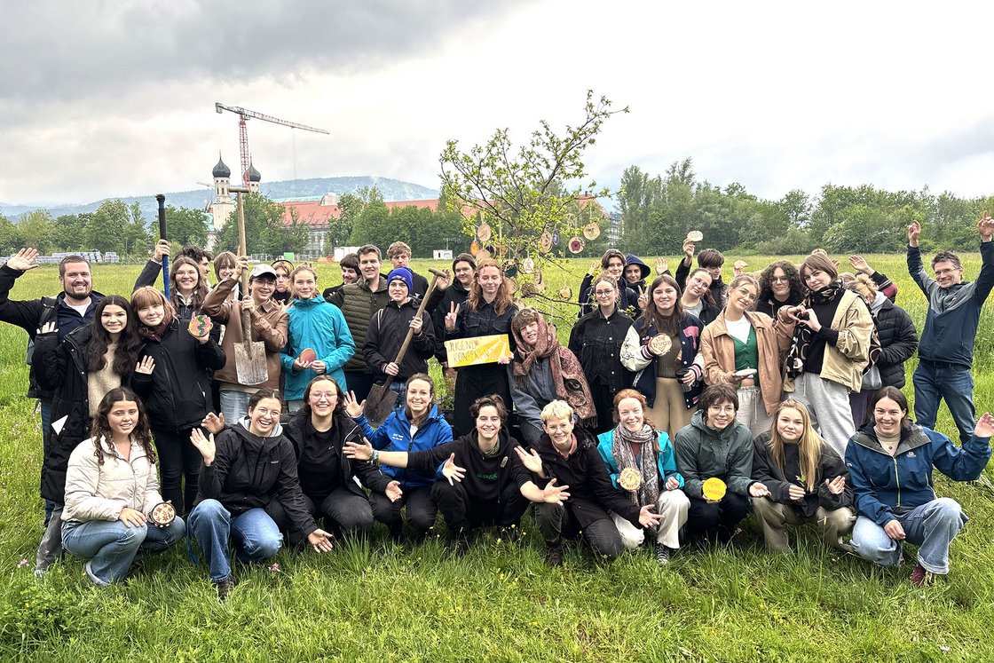 Eine vielfältige Gruppe von Menschen posiert auf einer Wiese mit einem geschmückten Baum und Werkzeugen. Kloster und Kran im Hintergrund.