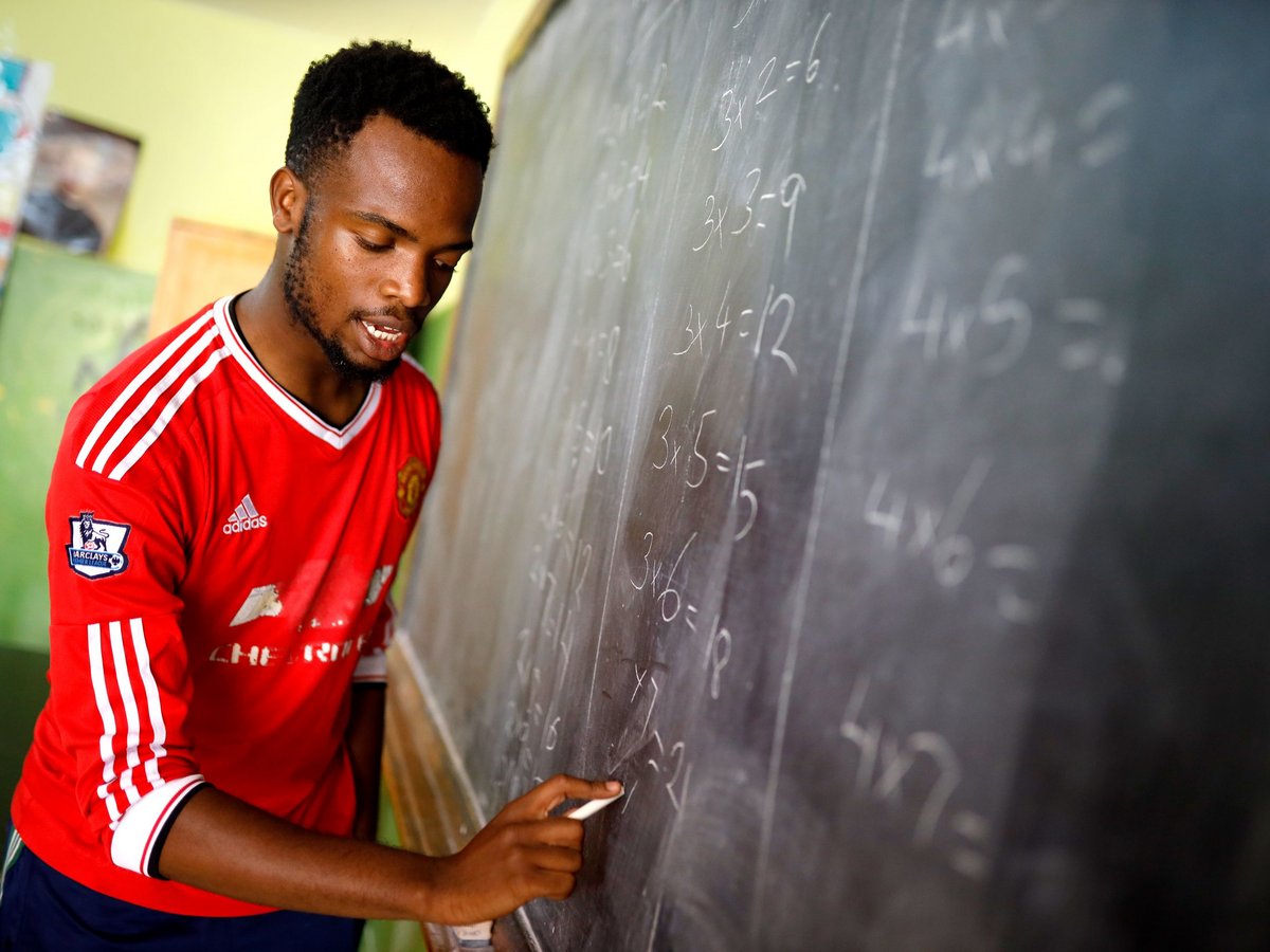 Junger Mann im roten Trikot schreibt Mathe-Aufgaben mit Kreide an eine Tafel, auf die Gleichungen konzentriert.