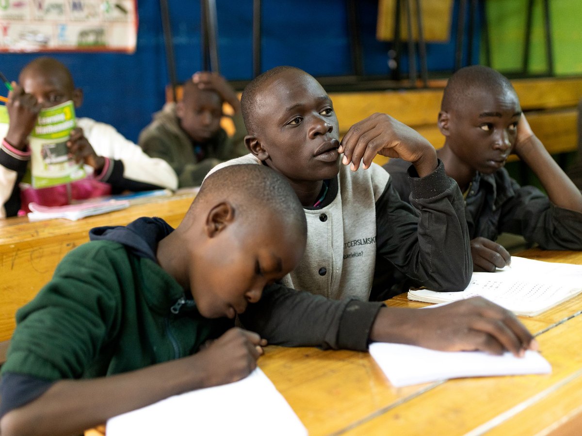 Mehrere Jungen in einem Klassenzimmer. Einer lehnt sich schreibend über sein Heft, ein anderer blickt nachdenklich auf. Lernsituation.